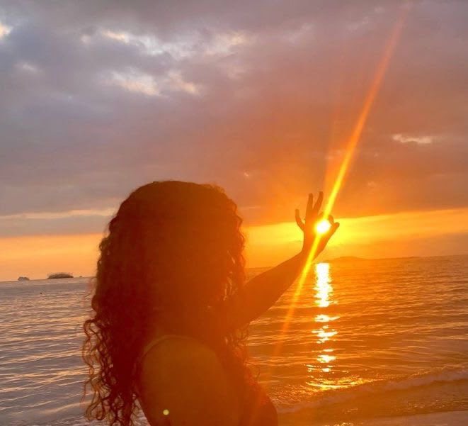 The image features a woman with curly hair standing near the water at sunset, holding her hand up towards the sun as if she's trying to capture its rays or simply enjoying the view. The setting sun casts a warm glow over the scene, creating an atmosphere of tranquility and beauty. The photo is taken from behind the woman, making it appear like she's looking out into the water at sunset.