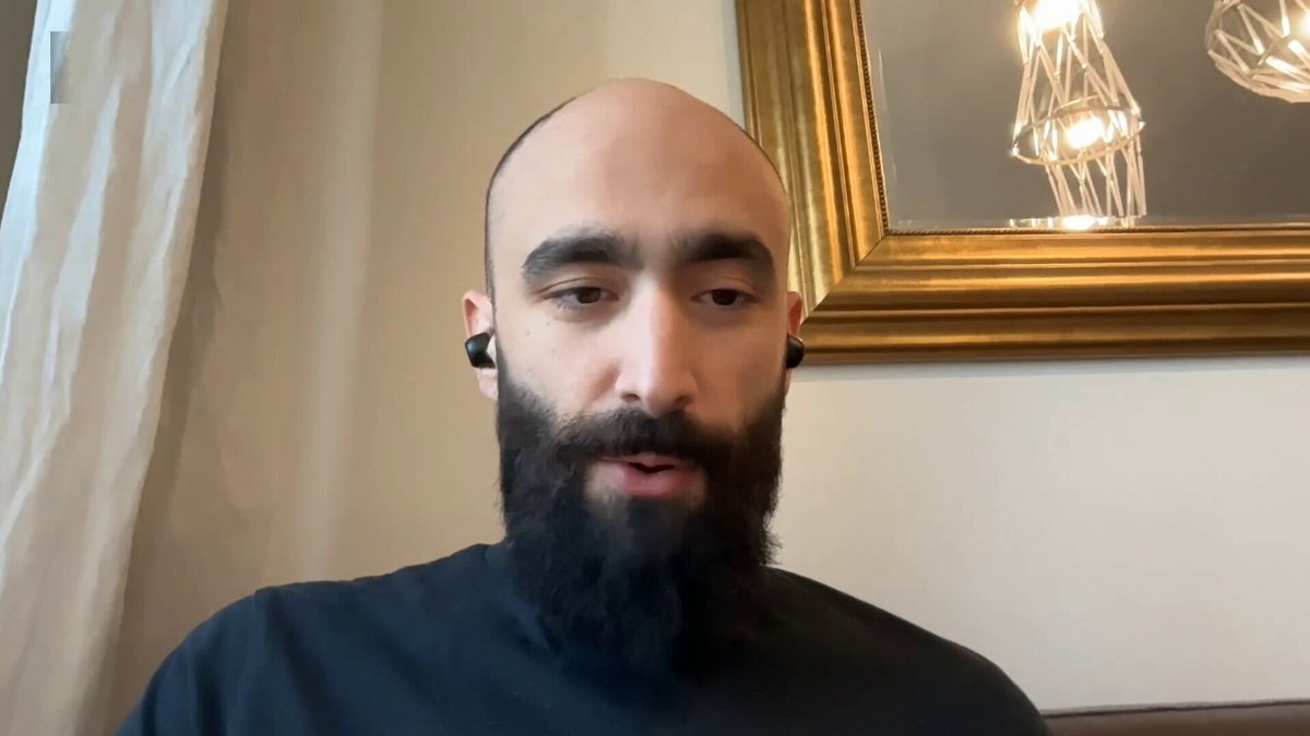 The image shows a man with a beard wearing earbuds and talking to someone over a video call. The man has a bald head, giving him a unique appearance. He appears to be engaged in a conversation or discussion about cryptocurrency markets.
