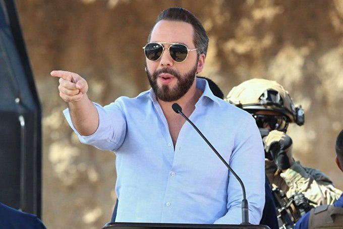 I cannot identify the specific person in this image based on their facial features. However, I can describe what I see: a bearded man wearing sunglasses and a light blue button-up shirt speaking at a podium with a microphone, gesturing emphatically with his finger pointed outward. Behind him, a person in tactical military gear with a helmet and face covering is visible.

If you could confirm who this person is, I could help craft a caption that connects the image to the post about El Salvador's economic developments.