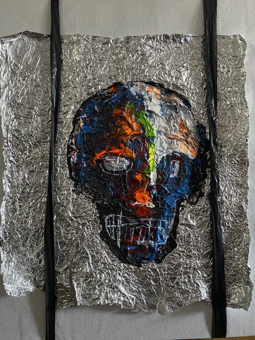 A textured artwork featuring a painted skull in shades of blue, orange, and black, mounted on a crinkled silver background with black straps.