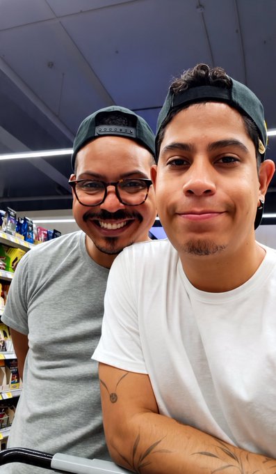 Two smiling men wearing caps taking a selfie in what appears to be a store aisle, with the man in front wearing a white shirt and sporting arm tattoos.