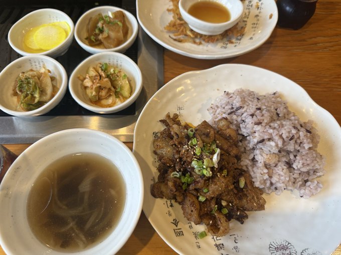A traditional Korean meal featuring grilled beef with purple rice on a decorative plate, accompanied by a bowl of clear broth soup, several small banchan side dishes, and dipping sauces.