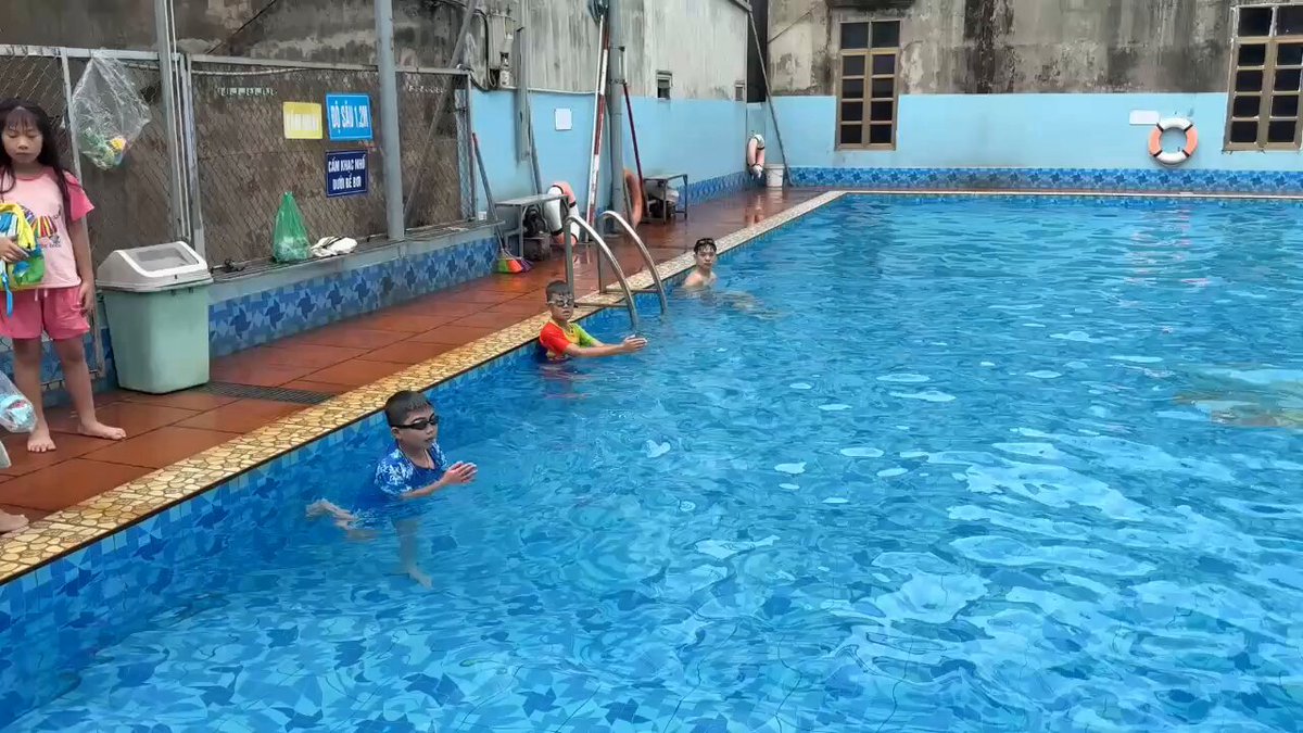 The image shows a group of people swimming in an indoor pool with blue water. There are at least six individuals in the pool, some wearing goggles while others do not. The scene appears to be taking place during the day under natural light. A child is learning to swim and is being assisted by two adults who are standing on the edge of the pool. The image also contains a caption that reads "The new project @CNPYNetwork and @quipnetwork is always facing difficulties like a child learning to swim but once the skill is mastered, joy and praise will come Success to you @quipnetwork @CNPYNetwork".
