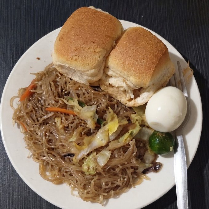 A white plate holds a meal consisting of stir-fried glass noodles with vegetables, two steamed buns, a boiled egg, and a small green citrus fruit.