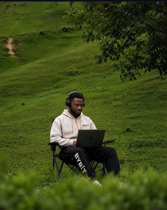 A man in a "BYBIT" hoodie and sweatpants sits in a camping chair on a grassy hillside, wearing headphones and working on a laptop—literally "touching grass" while embracing the crypto lifestyle with a relaxed "GM" vibe.
