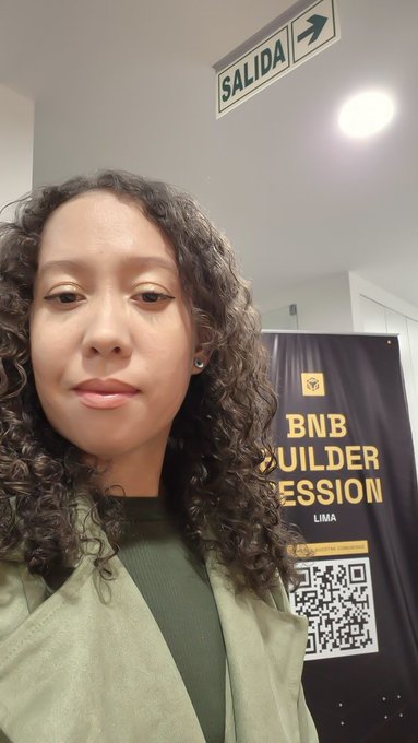 A selfie of a woman with curly hair wearing a green jacket, standing indoors beneath a 'SALIDA' exit sign. Behind her is a black banner for the 'BNB BUILDER SESSION' in Lima featuring a QR code.