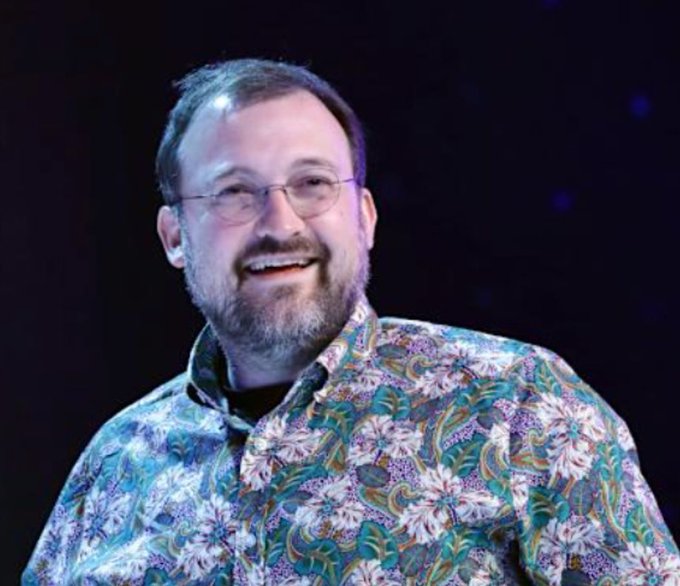 The image features a man wearing glasses and a colorful shirt, smiling at the camera with his hands on his hips. The background of the photo appears to be dark or black, which contrasts with the bright colors of the man's shirt. There is also text in the image that reads "CHARLES HOSKINSON WARNS MORE $ADA PROJECTS ARE GOING TO DIE! Another tough day for Cardano, following TapTools shutting down. Hoskinson said it will be a tough year for the project". The man's facial expression and body language suggest he is confident or proud of his work on the projects, despite the challenges they may face.
