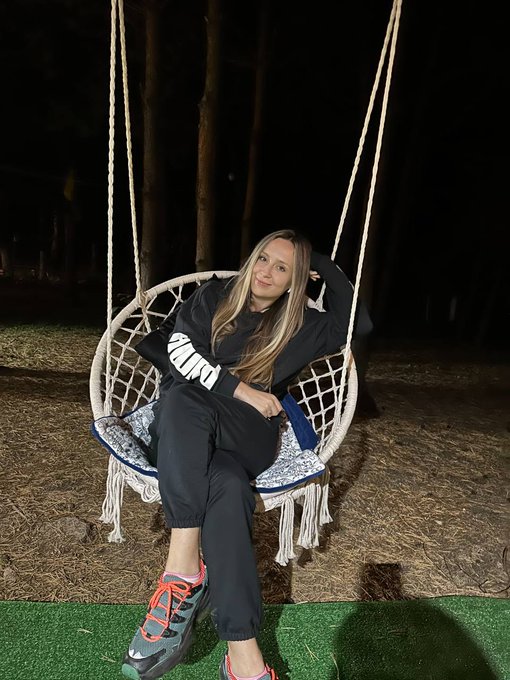 A nighttime photograph of a young woman with long blonde hair sitting in a large, round macrame hanging swing, wearing black sweatpants and a black sweatshirt with white sleeve stripes.