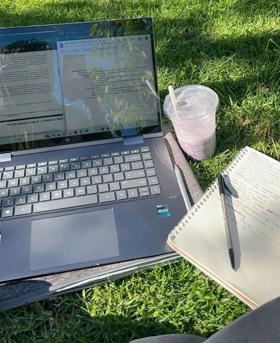 The image shows a laptop computer sitting on top of a notebook, with a drink placed next to them. The scene takes place outdoors, possibly on grass or dirt, giving an impression of a casual work environment. The focus of the image seems to be on the user's ability to manage their digital assets and access multiple accounts across chains without fragmentation issues. This is highlighted by the presence of the self-custodial account feature in the cryptocurrency space.