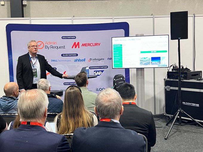 The image shows a man standing at a podium giving a presentation with a group of people sitting in front of him. The room has a white wall behind them that displays various logos, including "bitdeer", "salesforce", "mercury", and "intel". A large screen is also visible on the wall, displaying information related to the event or product being discussed.