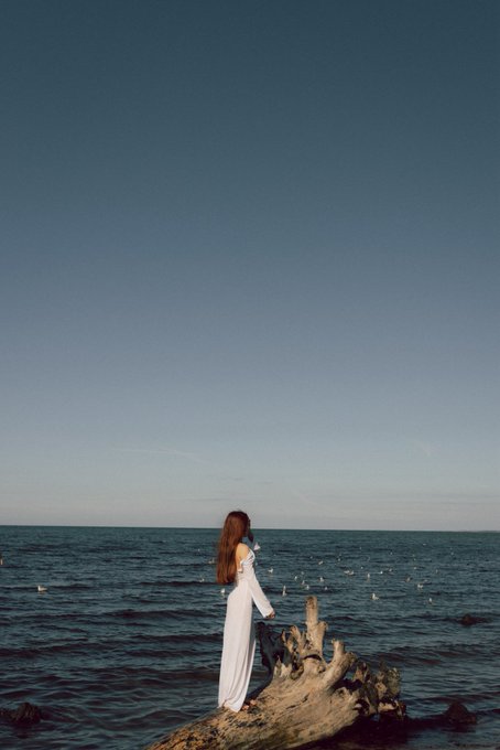 A woman in a white dress stands on driftwood by the sea, facing the ocean under a clear blue sky.