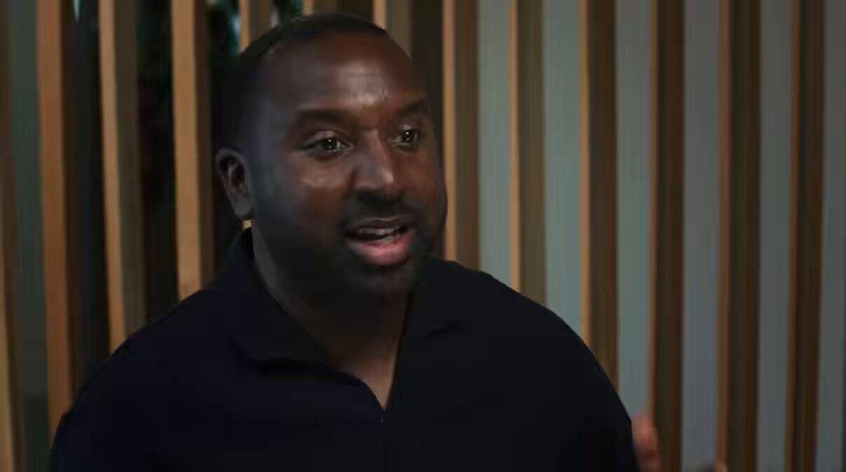 The image shows a man wearing a black shirt with his face partially visible as he looks at someone or something off-camera. The background features vertical wooden slats, which could be part of a wall or a door frame. There are no other objects in the image besides the man and the wooden slats.