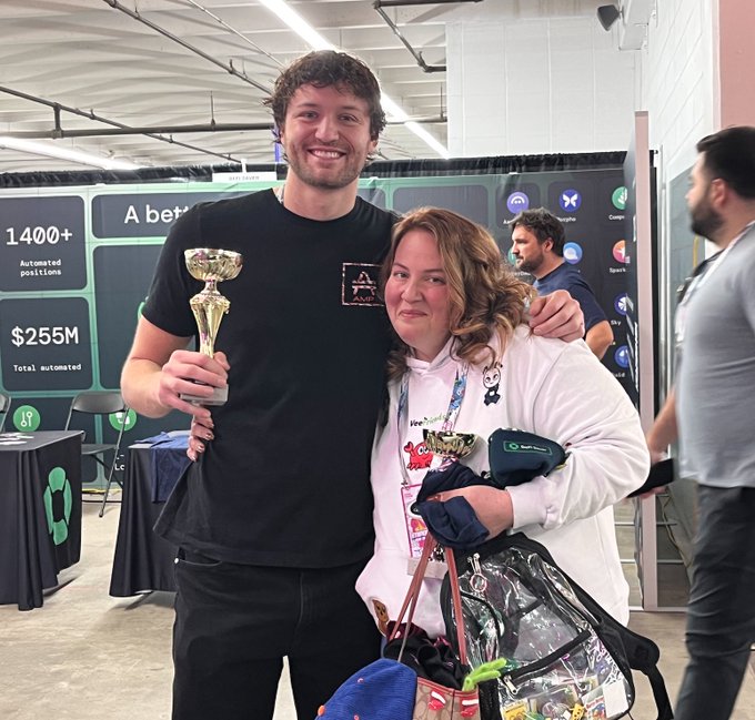 A smiling young man in a black 'AOAI' t-shirt holds a gold trophy while posing with a woman in a white hoodie at what appears to be a convention, with a booth displaying '1400+ Automated positions' and '$255M Total automated' behind them.