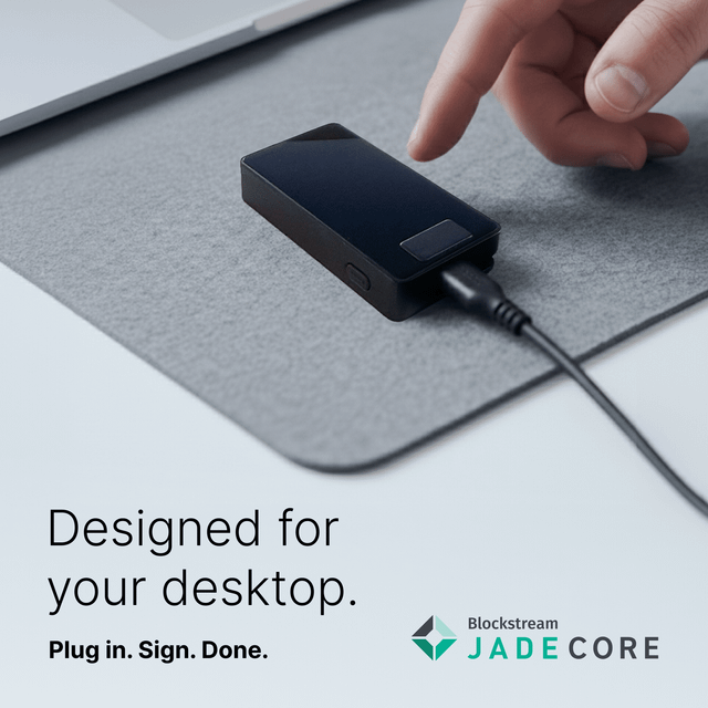 The image shows a person's hand pointing at a small black device that appears to be a USB drive or flash drive, placed on top of a gray mouse pad. The background of the image is white with some text visible, which reads "Designed for your desktop. Plug in Sign. Do." and "Blackscore JadeCore". This suggests that the person might have recently plugged in their device to charge it, as indicated by the phrase "blackscore jadecore" in the image description.