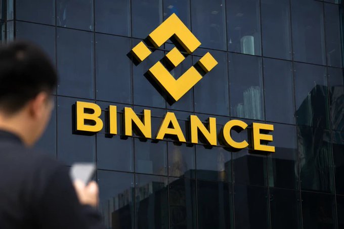 A person looks at a smartphone in the foreground while the yellow 'BINANCE' logo is displayed on a building's glass facade behind them.