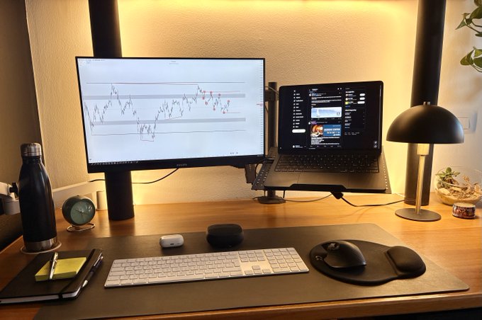 A neatly organized desk setup featuring a widescreen monitor displaying stock charts alongside a laptop, with the caption "GM Friends!" suggesting a morning greeting shared by a trader or investor to their online community. The workstation includes an Apple Magic Keyboard and trackpad, wireless mouse, AirPods case, water bottle, and warm ambient lighting that creates an inviting atmosphere for analyzing the markets.