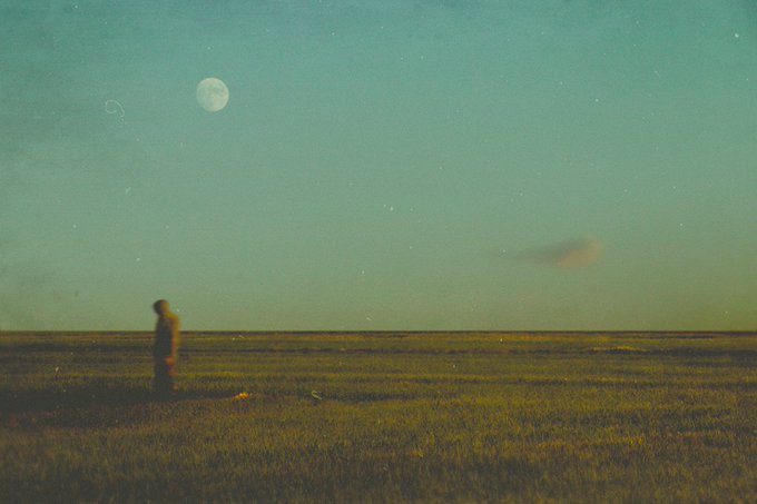 A moody, grainy photograph captures a solitary figure standing in a vast grassy plain beneath a pale green sky featuring a moon and a faint ringed planet.