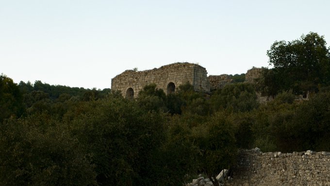 An ancient stone ruin with arched openings, surrounded by lush green vegetation and olive trees under a pale sky.