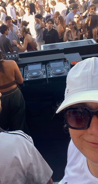 A selfie of a woman wearing a white cap and sunglasses at a DJ booth with a crowd in the background.