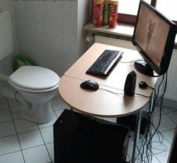 A bathroom doubles as a computer workstation, with a toilet beside a desk holding a keyboard, mouse, monitor, and speakers; multiple cables hang beneath the desk.