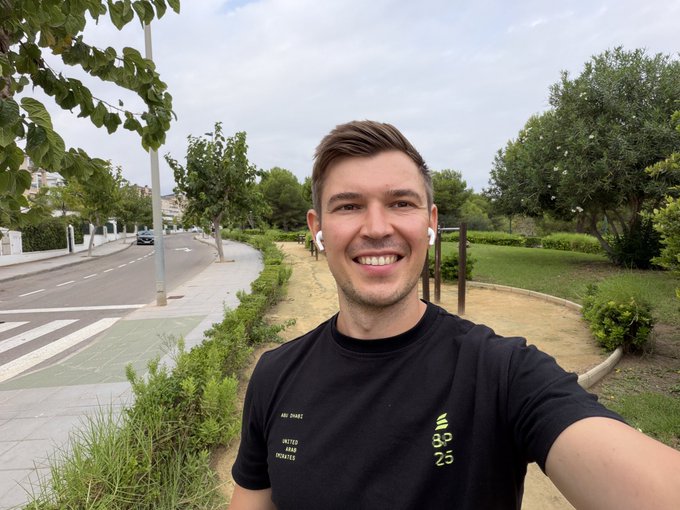 A smiling man wearing a black athletic t-shirt (with 'UNITED AIR 2024' and numbers '33 23') and white earbuds takes a selfie outdoors on a tree-lined street with a bike lane.