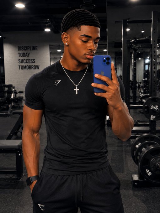 A muscular young man with braided hair taking a mirror selfie in a gym while wearing a black athletic shirt with a cross necklace, with the motivational text 'DISCIPLINE TODAY SUCCESS TOMORROW' visible on the wall.