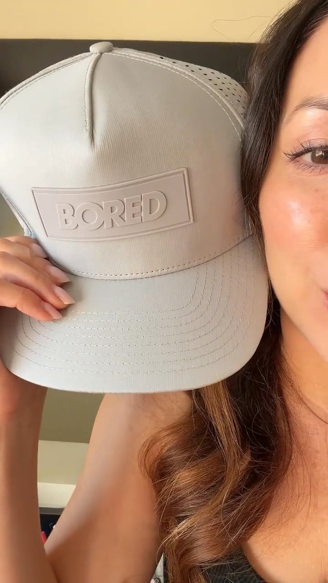 A woman holds up a light gray baseball cap with the word 'BORED' embossed on a rectangular patch, positioned next to her face.