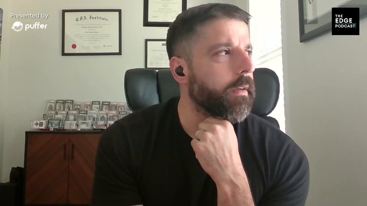 The image shows a man wearing glasses, sitting at his desk with his hand placed over his chin as if deep in thought or contemplation. The background features a white wall with several framed pictures hanging above him. There are also multiple books scattered around the room, suggesting that he might be working on some research or studying for an exam.