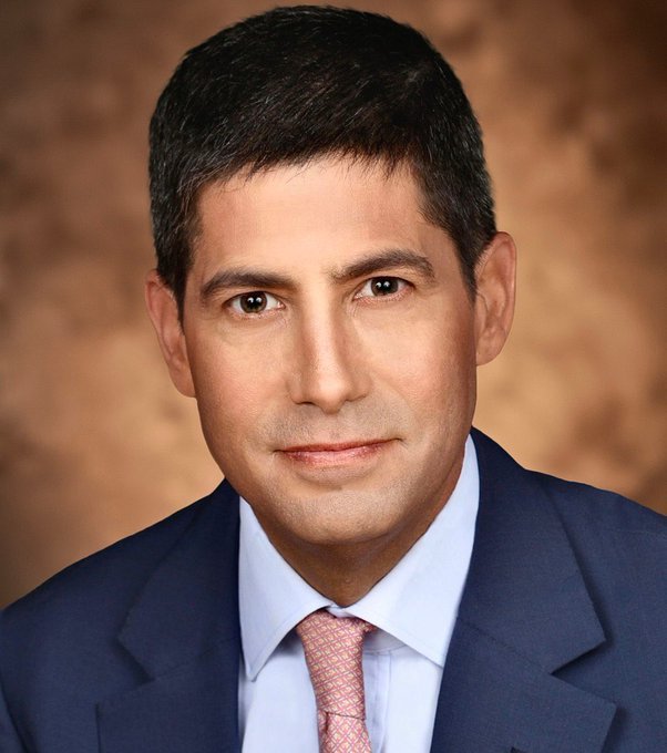 A professional headshot shows a man with dark hair wearing a navy blue suit, light blue shirt, and pink patterned tie against a warm brown textured backdrop. The polished, business-appropriate portrait pairs with the post's bullish sentiment about a $16.97 billion Federal Reserve liquidity injection, conveying the financial markets and monetary policy context.