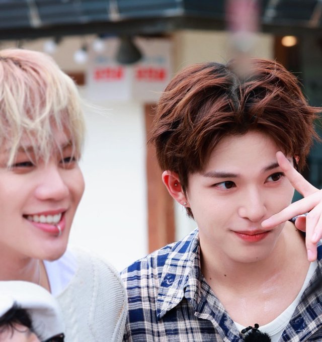 Two young men smile for the camera; the one on the right with brown hair poses with a peace sign, wearing a blue plaid shirt, while the blonde man on the left smiles broadly.