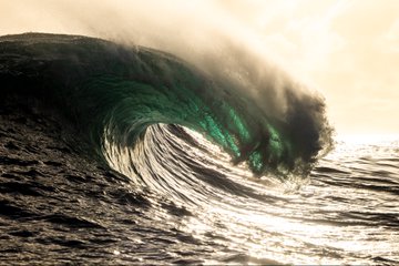 A large ocean wave curling with green-tinted water and misty spray against a sunset sky.