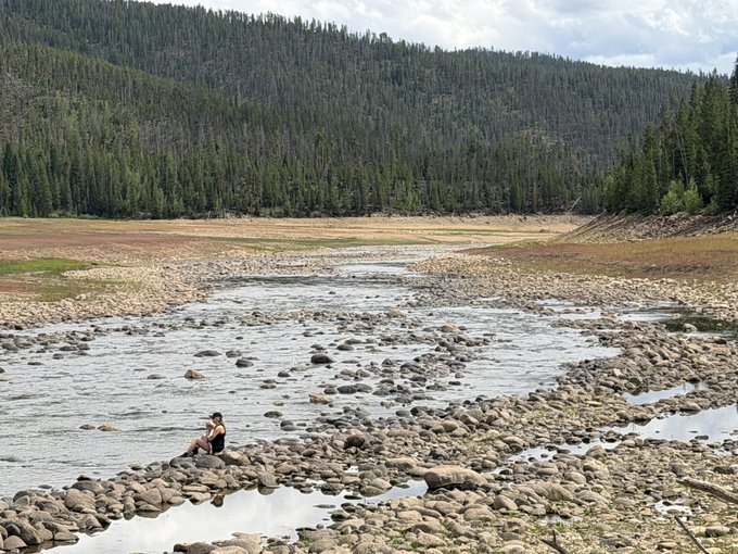 A scenic landscape showing a rocky riverbed with shallow water, where a person sits on the stones, surrounded by forested mountains under a partly cloudy sky.