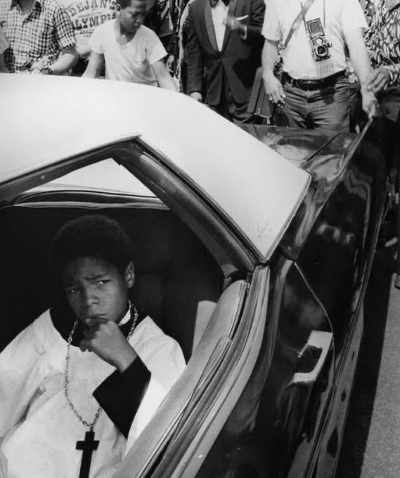 A young boy wearing a cross necklace sits pensively in the back of a car, surrounded by onlookers and a photographer, with the partial text "DIAN'S OLYMPI" visible on a jacket in the background. The image's contemplative tone seems to contrast with the celebratory post encouraging friends of "Dumex" to "win today."