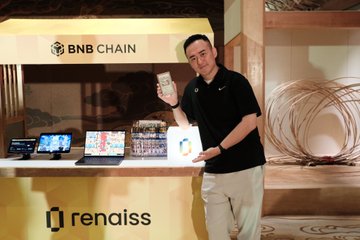 A man holding a smartphone at a promotional event displaying 'BNB CHAIN' and 'renaiss' branding, with several laptops set up on the counter behind him.