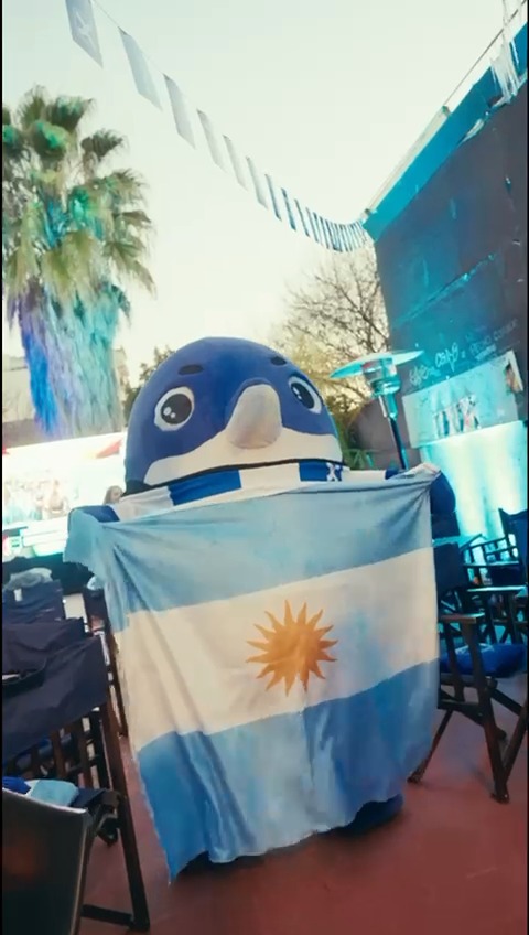 A BingX mascot with large expressive eyes holds the Argentine flag with its sun emblem at an outdoor live screening event in Buenos Aires, matching the post about the community celebrating the match with unmatched vibes.