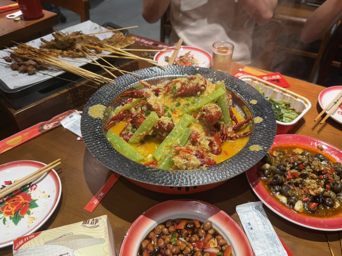 A spread of Chinese dishes on a dining table, featuring a central bowl of crab in a creamy orange sauce with green vegetables, surrounded by skewers, peanuts, snails, and other side dishes.