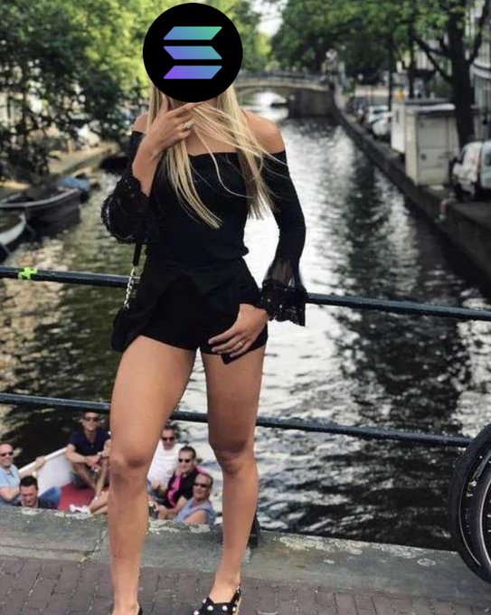 A person in a black off-shoulder top and shorts stands on a bridge over a canal with boats and crowds below, their face obscured by a Solana logo.