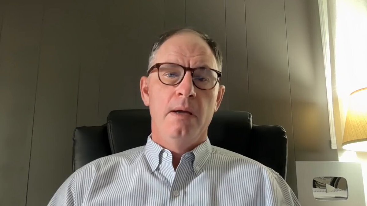 The image shows a man wearing glasses sitting in an office chair, looking at the camera with a serious expression. He appears to be engaged in a video call or a live stream, as indicated by the presence of a laptop computer placed on his lap. The background features a wall and a window, suggesting that he is likely working from home or in a professional setting where remote communication is common.