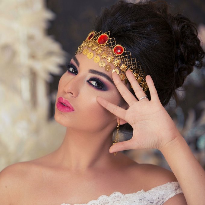 A beauty portrait of a woman with dark hair styled up, wearing a gold headpiece with red jewels, dramatic eye makeup, and bright pink lipstick, posing with her hand near her face.