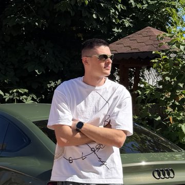 A young man in a white graphic t-shirt and sunglasses stands with crossed arms in front of a green Audi car and lush greenery on a sunny day.