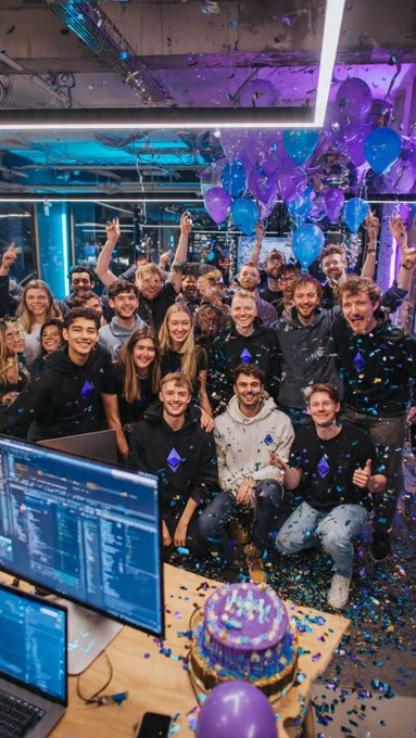 A large group of young people celebrating in an office space with blue and purple balloons, confetti falling, and a cake on a desk in the foreground.