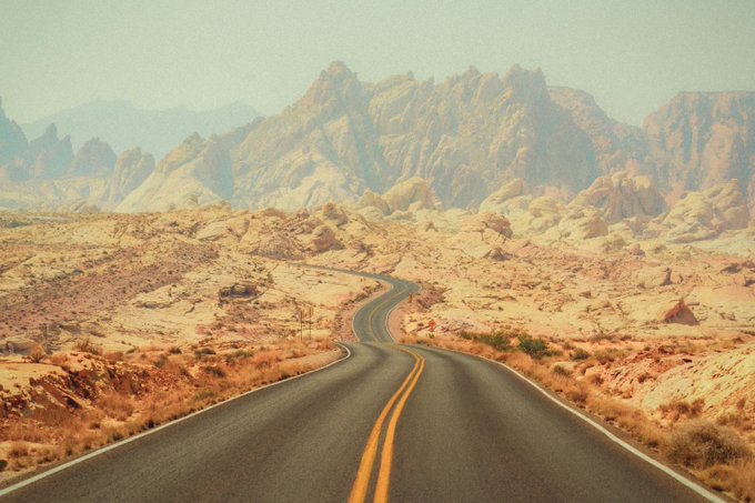 A desert landscape photograph featuring a winding paved road with yellow lines leading toward rugged, reddish-brown rock formations and mountains under a hazy sky.