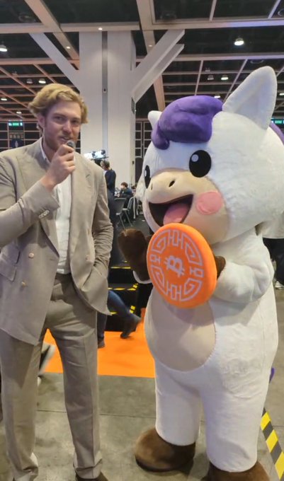 A well-dressed man in a beige suit smiles and gestures near his chin while posing beside a large white and purple cow mascot holding an orange "Bitcoin Pizza" token, teasing an upcoming interview. The orange Bitcoin-themed sign and cryptocurrency mascot hint that the interview may focus on Bitcoin or crypto-related topics.