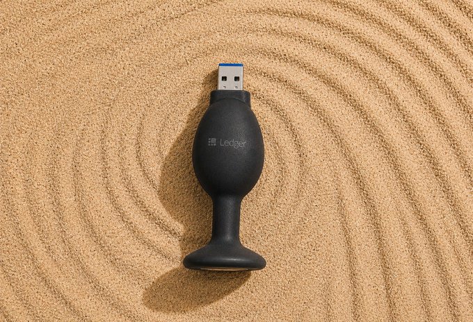 A black Ledger hardware cryptocurrency wallet with a blue USB connector, resting on sand with concentric circular raked patterns around it.