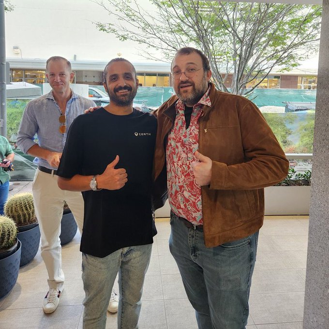 Two men pose together smiling and giving thumbs up, with a third man in the background, in what appears to be a shopping mall or plaza.