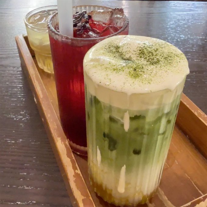 A wooden tray holding three beverages: a tall glass of iced red drink with a straw, a small glass of yellow liquid, and a layered iced green matcha drink topped with creamy foam and matcha powder.
