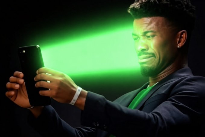 A man in a dark suit looking at a smartphone with a displeased expression, illuminated by a bright green glow reflecting off his face and the phone against a dark background.