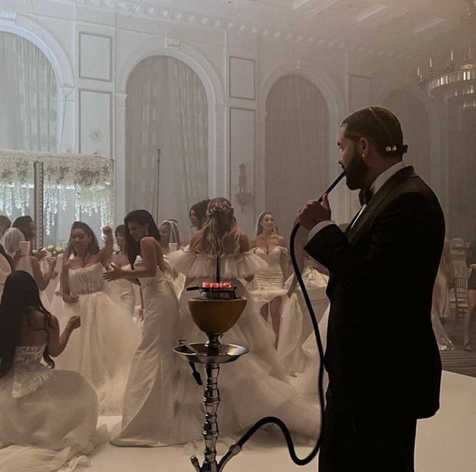 A scene showing a man in a dark suit smoking a hookah while several women in elegant white gowns pose and take photos in a grand, ornately decorated hall.