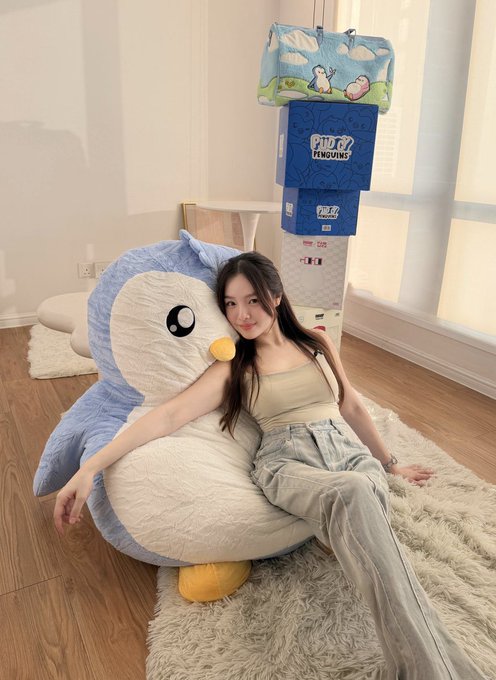 A woman reclining against a large Piplup plushie on a fluffy rug, with stacked FWZ (FunZow) branded boxes visible in the background near a window.