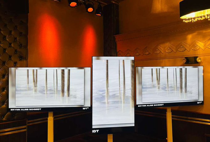 Three monitors in a dimly lit, ornately decorated room with red lighting display artwork by Bryn Alise Schmidt, showing vertical wooden poles standing in a misty landscape.